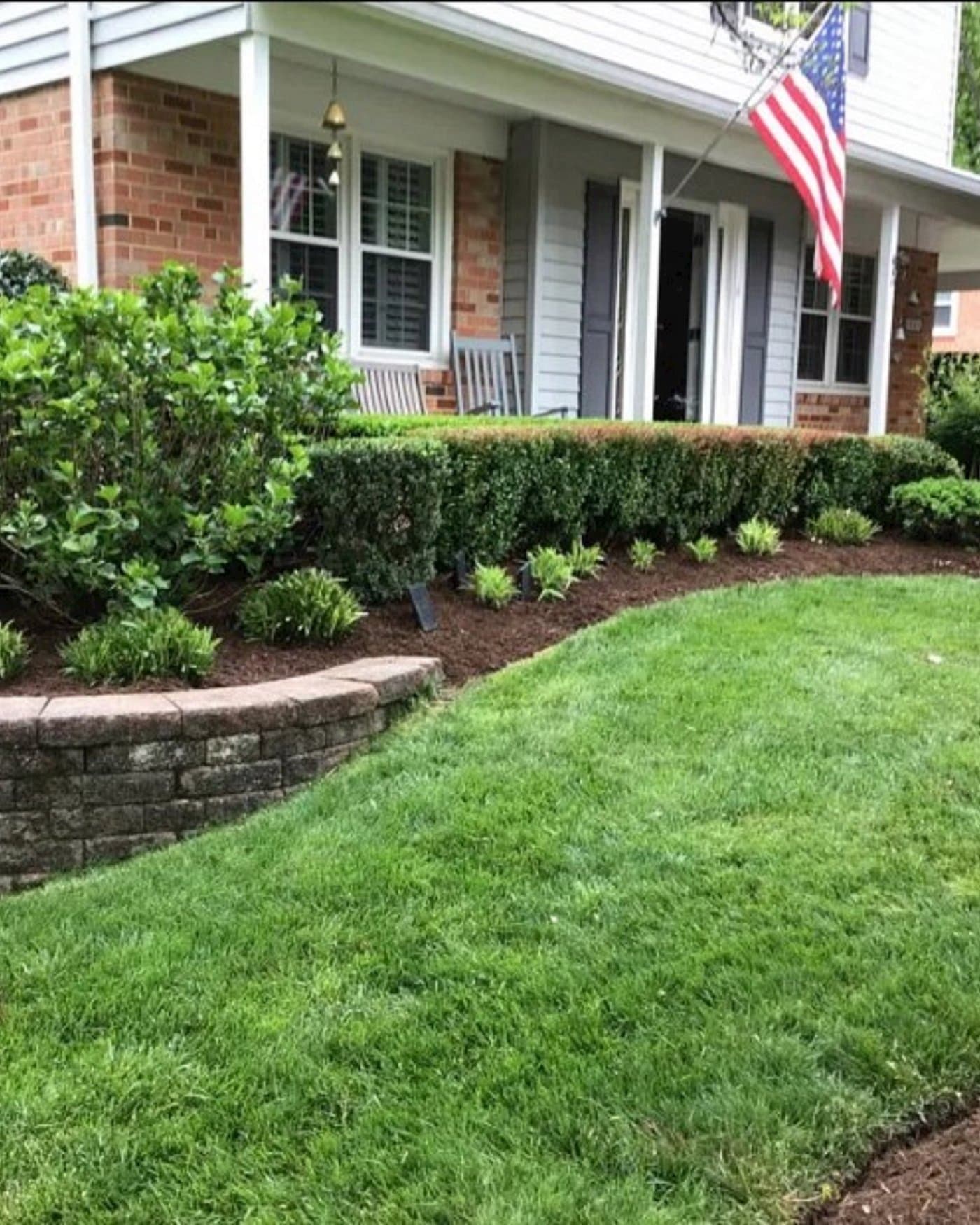 A two-story brick home with a covered porch and American flag, fronted by a trimmed hedge, mulched garden bed, stone retaining wall, and tidy lawn